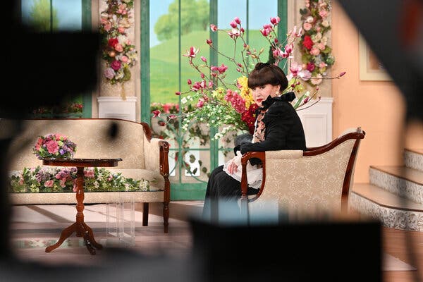 A woman sits on a chair in what appears to be a living room decorated with flowers and a view of a green hill outside.